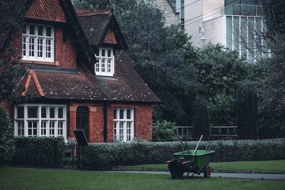 A red brick house with a pitched tiled roof featuring white-framed double-glazed windows and a central black front door, set within a well-maintained garden. The garden includes neatly trimmed hedges