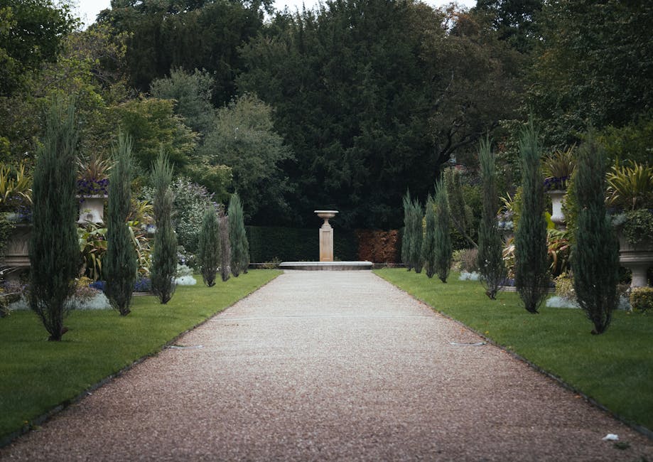 A straight gravel pathway runs through a well-maintained garden with neatly trimmed green grass on both sides. Tall, slender cypress trees line the edges of the path, each planted in large decorative 